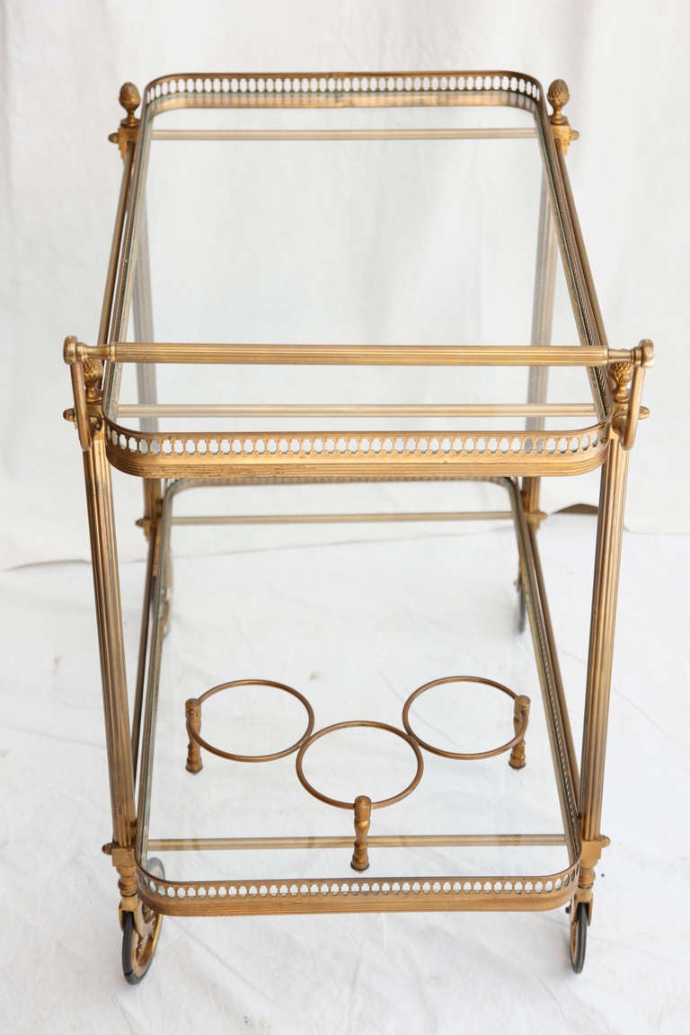 This elegant bronze bar cart features Louis XVI style details of fluted columns and pinecone finials. With smoothly rolling bronze framed wheels, two ample glass shelves, and a built-in bottle holder, this piece will function beautifully for serving