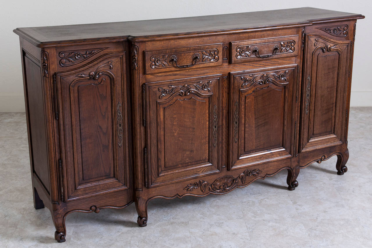 The delicately carved Louis XV motifs of this enfilade interact beautifully with the deep heart grain of its solid French oak body. With four cabinet doors and two drawers, this piece will provide ample storage for a large dining space. Its unusual