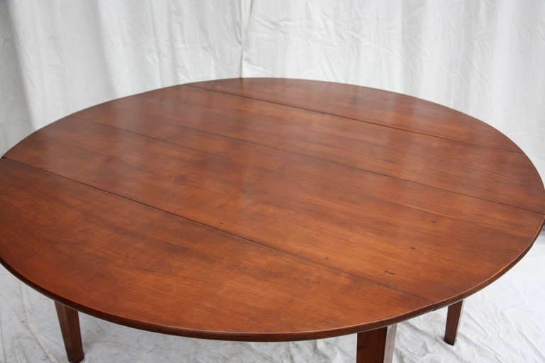 This c. 1900 artisan made large round dining table from Normandy, France was made from thirteen inch wide cherry wood planks.  Its hand pegged legs provide a sturdy base, and its two drop leaves allow it to be placed flush against a wall or to pass