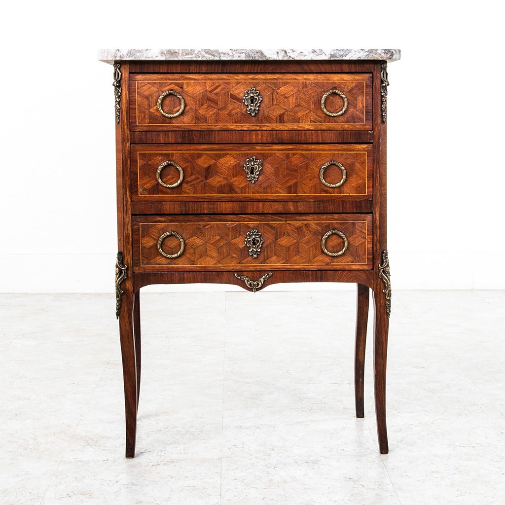 This elegant petite Louis XV - Louis XVI transitional style chest of drawers features geometric parquetry, bronze hardware, and a light grey marble top. The slender cabriole legs and ormolu belie its Louis XV characteristics, making this a great