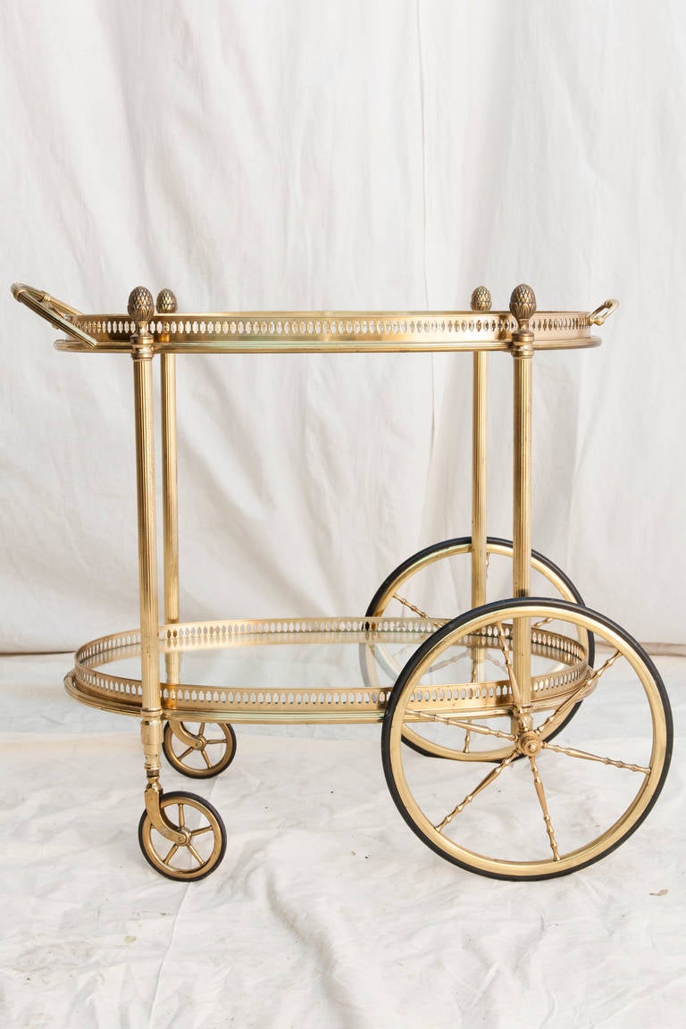 This mid twentieth century oval brass bar cart from France features a brass gallery around each of its glass shelves.  Its top shelf is a removable tray, and its four legs are capped with Louis XVI pine cone finials.   An elegant and charming piece