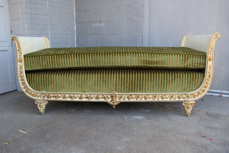 This charming iron daybed has an antiqued cream enamel finish with gilt raised floral detail and a central Putti.  The pristine and comfortable forest green velvet upholstery with gold stitched striping coordinates beautifully with the finish .