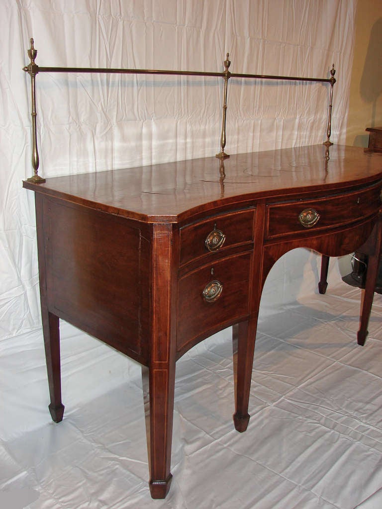 This graceful, period Georgian, mahogany serpentine sideboard has a cross-banded top with stringing and is further enhanced by a brass rail. There are brass, oval, bale pulls with lion masks. The left side has a door, while the right side drawer is