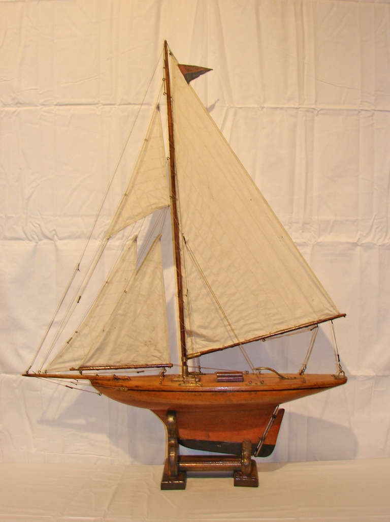 Handsome wood pond yacht with stitched canvas sails and brass fittings. Incised line in wood defines the gunwale. Ensign colors have aged to a muted red and blue. The yacht rests on a wood stand.