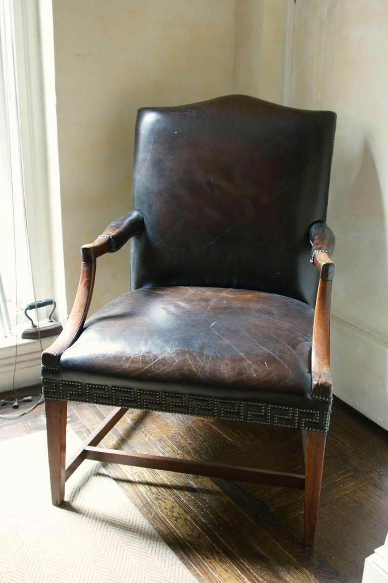 This is a mahogany Gainsborough chair of graceful proportions. The chair has a curved upper back and curved arms with antiqued brass nailhead trim around the dark brown leather armrests. A boldly distinctive border of nail heads in a Greek key