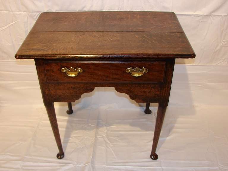 This early Georgian oak side table has a two-part top, shaped apron and sides and tapering legs terminating in pad feet. The bale style brass pulls on the drawer appear to be original. It would make a great lamp table.