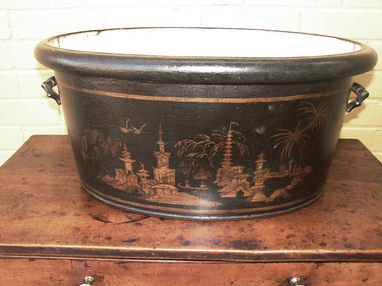 This black tole footbath is beautifully painted with a gilded chinoiserie design of a landscape with palm trees, pagodas, figures rowing in a boat and birds flying above. The footbath has shaped metal handles with a dark patina and an ivory painted