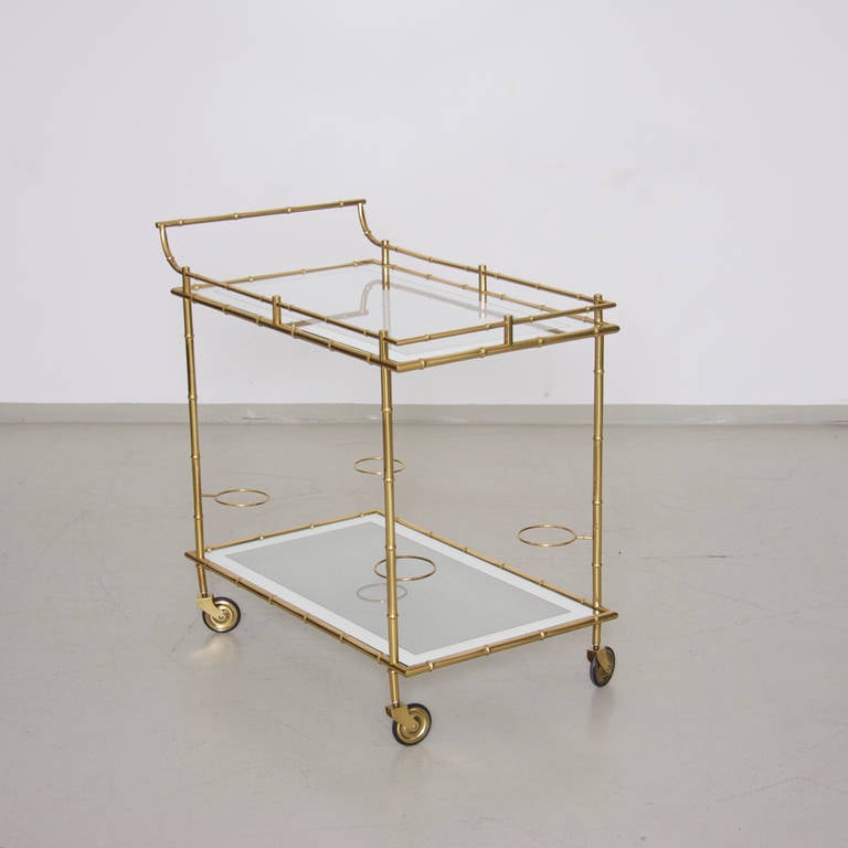A midcentury brass bar cart in faux bamboo style from Maison Bagues. The cart features two clear glass tops with chrome galleried edges and four bottle holders.