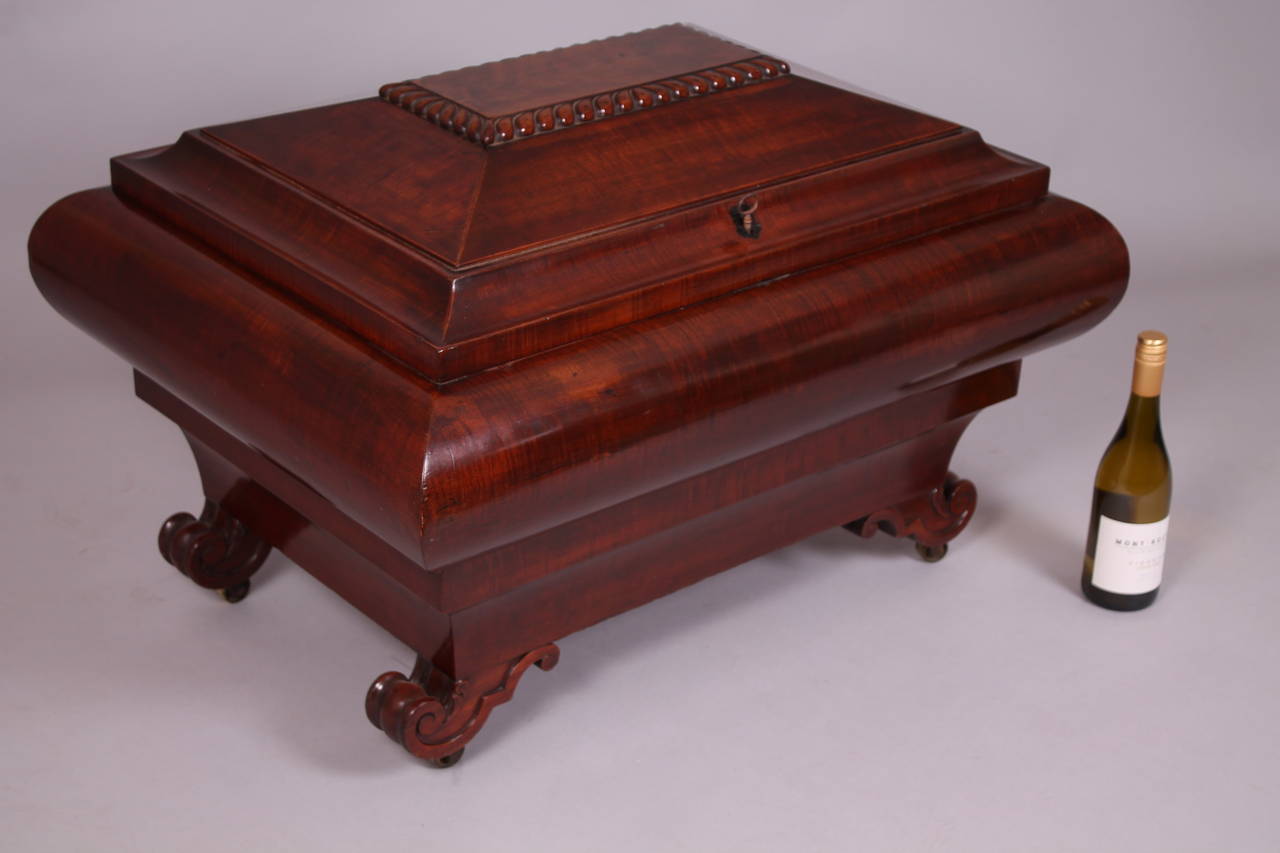 A monumental and very important mahogany wine-cooler of fine quality; of bold sarcophagus form with bombe sides and a domed and gadrooned lid, on carved volute feet; the lead-lined interior divided into two sections for six and twelve bottles;