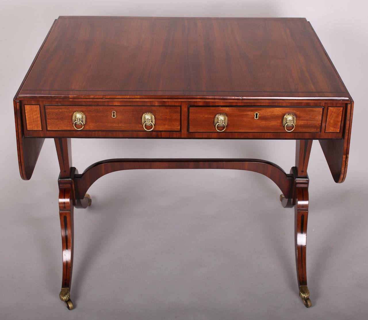 Regency period mahogany sofa-table inlaid with ebony bandings and decorative motifs; the frieze fitted on each side with one real and one dummy drawer with original brass sphinx's head ring-handles, on an elegant base with tapered end-supports, an