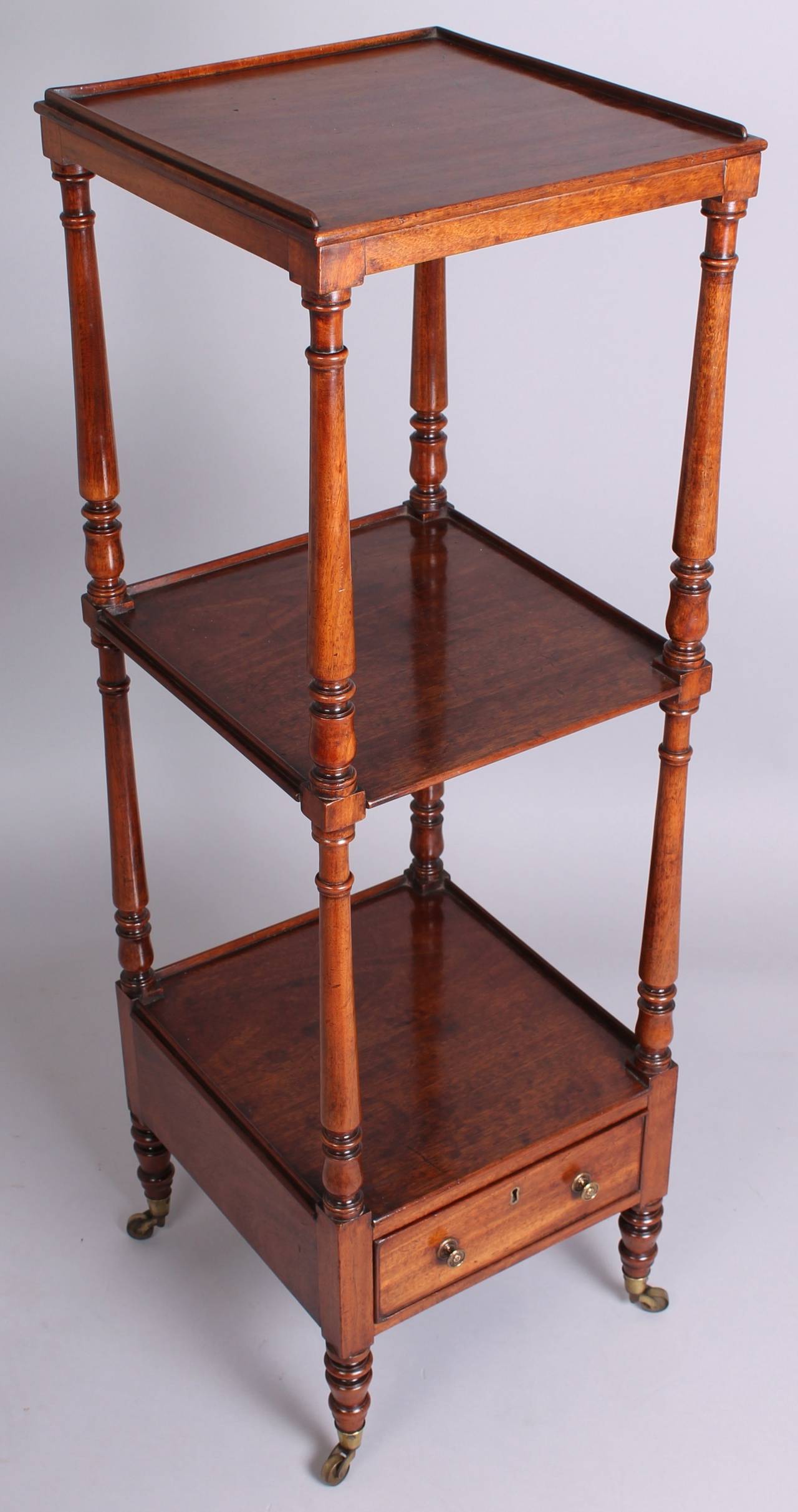 A late George III period mahogany small whatnot; the almost-square galleried shelves mounted on turned baluster-shaped supports and legs with brass cup-castors; the base fitted with a mahogany-lined drawer. Circa 1815