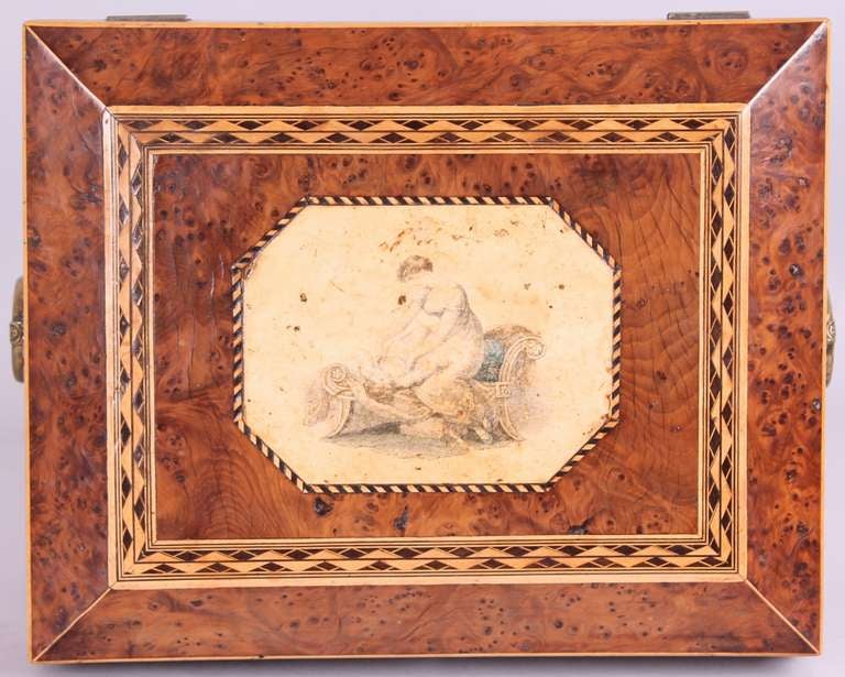 Regency period burr yew work-box; the top with chevron bandings and a coloured engraving in the style of Adam Buck; with brass ring-handles and paw feet; the interior with a lift-out tray with its original fittings and pink lining-paper. Circa 1815