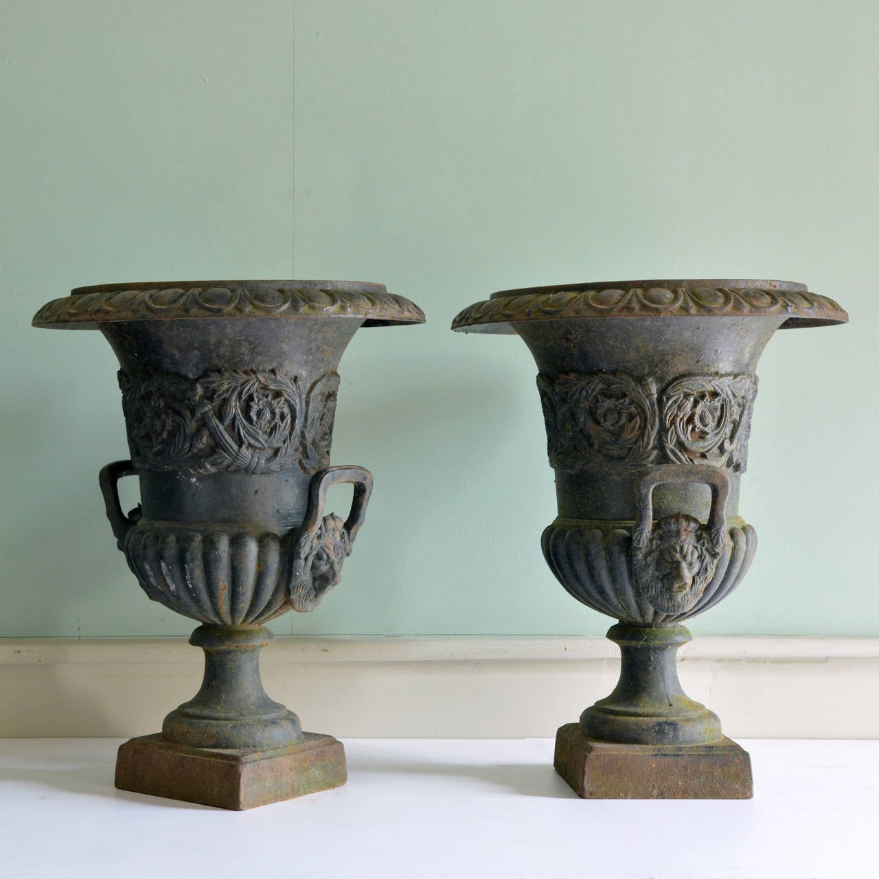 A pair of Victorian style cast iron urns, in the manner of the Handyside foundry,
the egg and dart moulded rims above body with typical foliate casting, the gadrooned bodies with lion mask handles on socle and square plinth bases. 

Available to