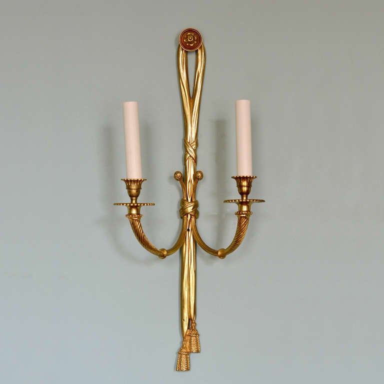 A pair of Louis XVI style brass wall sconces each backplate composed of draping with tasselled finials, two branch with decorative drip pans.

Available to view at Brunswick House.