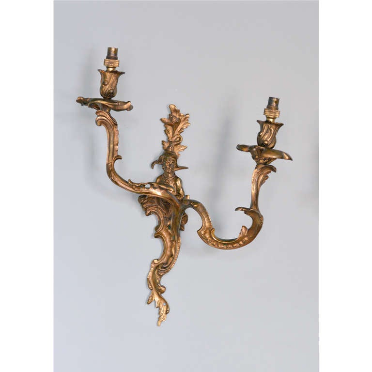 A pair of Louis XV style ormolu two branch wall lights, 19th century, each back-plate cast with sprouting foliage above Chinoiserie figure with c-scrolls and acanthus below, flanked by pierced c-scroll and acanthus branches, with foliate cast