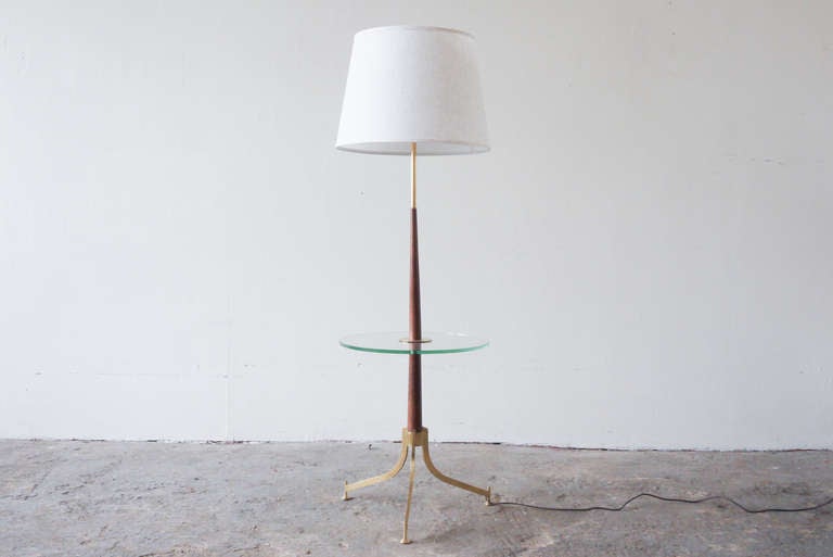 This Gerald Thurston floor lamp for Lightolier stands on three splayed brass legs. The stem tapers as it ascends, transitioning from wood to brass near the top. A circular glass table floats around the stem, making this lamp a multi-purpose utility.