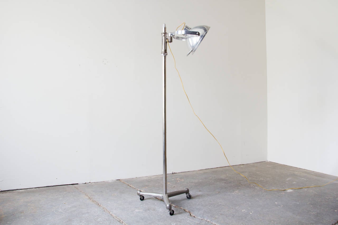 An adjustable medical floor lamp in steel by Prometheus. The head of the lamp can be moved anywhere along the vertical rod as well as tilted and swiveled on the joint. The lamp rolls on three casters.