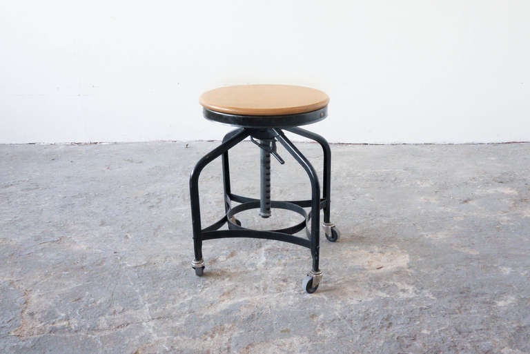 This antique Toledo drafting stool is adjustable to various heights. It has a circular wooden seat, atop a steel frame, and exhibits the classic Toledo ribbon thin rails. It rolls on four industrial casters.