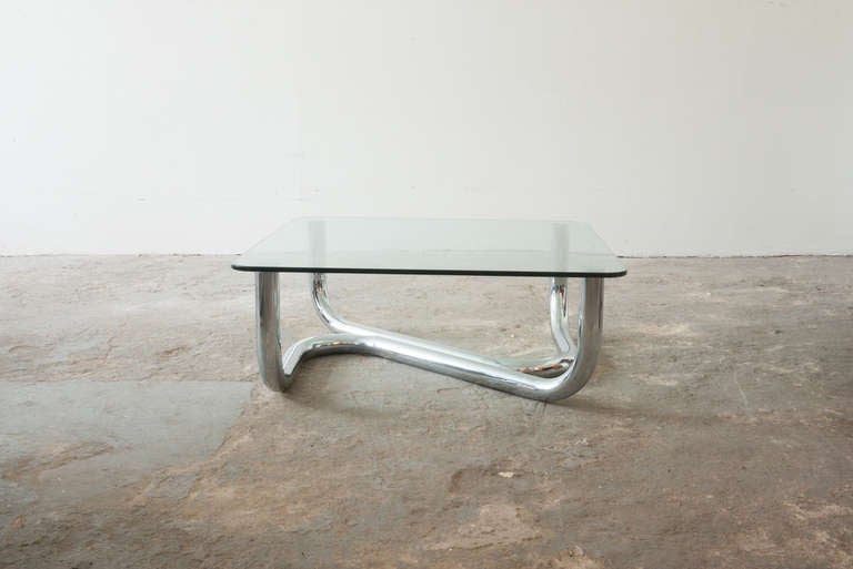 This coffee table, of chrome and glass, is elegantly curvaceous. Its tubular base has the effect of appearing to be in two parts, and organically separate as the tubes move vertically. The base supports a thick, substantial rounded square piece of