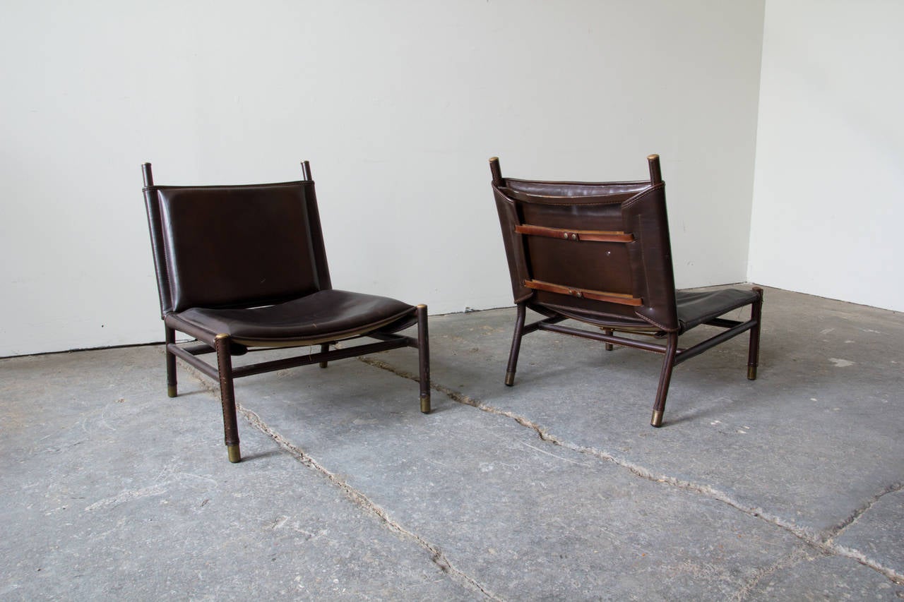 A pair of scarce slung slipper chairs by Jacques Quinet. The frames are comprised of tubular steel and are entirely wrapped in leatherette and saddle stitched. Brass sabots adorn each extremity. The seats and backs are foam, upholstered in