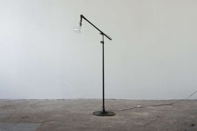 This floor lamp features a thick, substantial shade of hand blown glass. The body of the light is steel. The base is a thick industrial era gear, providing for sturdy use. The arm booms, and adjusts with a small gear clamp.