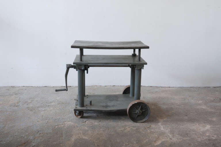 This machine-age cart raises and lowers via a crank. It is substantial in weight, and durably constructed. It rolls on four wheels, the larger of which are fixed in place.