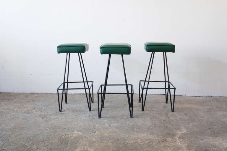 This set of three wrought-iron bar stools bear a resemblance to the hairpin stools reminiscent of Frederic Weinberg. Each angled leg flows from the top, meets the floor at the foot, and rises to support the foot rest. The seat cushions have been