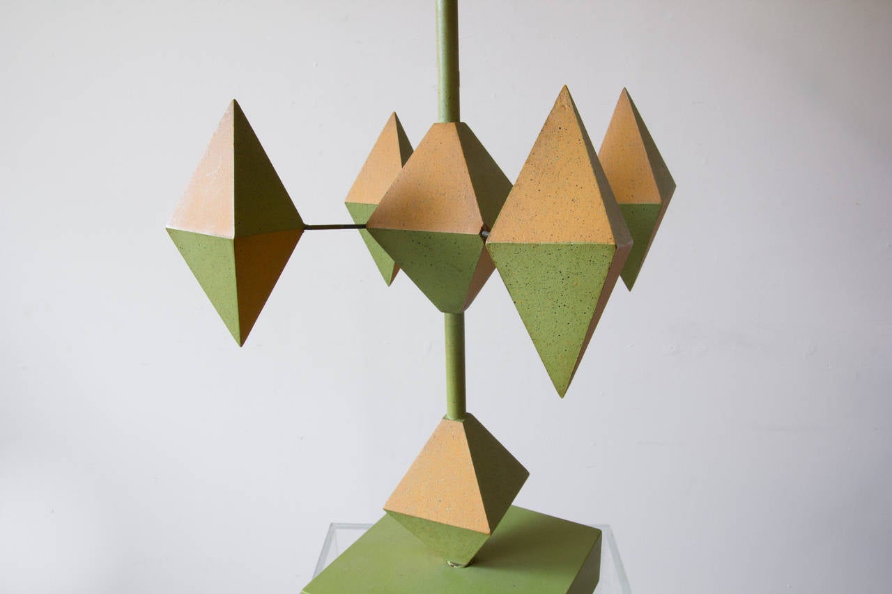 A unique geometrical table lamp comprised of painted wood and wire. The diamond shapes extruding from the center of the lamp, affixed by wire, are painted in complimentary hues of amber and green, creating a playful optical effect. 

Base