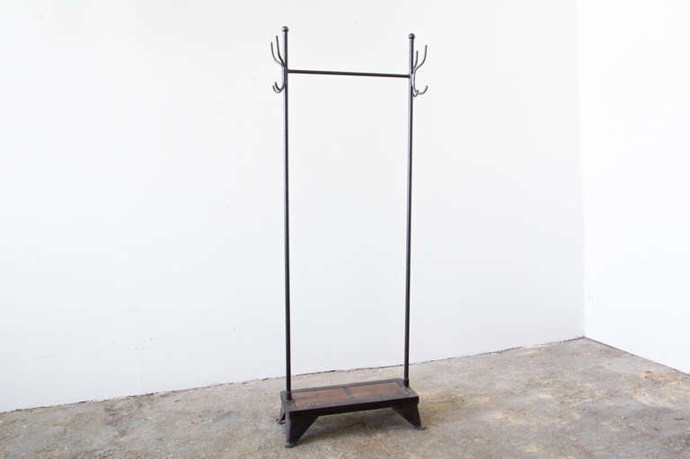 A slim coat rack of iron and wood. At the base, steel legs frame a reclaimed bleacher seat platform, which can be used for shoe storage. Two iron rods connect with a stretcher at the top, where hangers can be used to hold coats. Four hooks on each