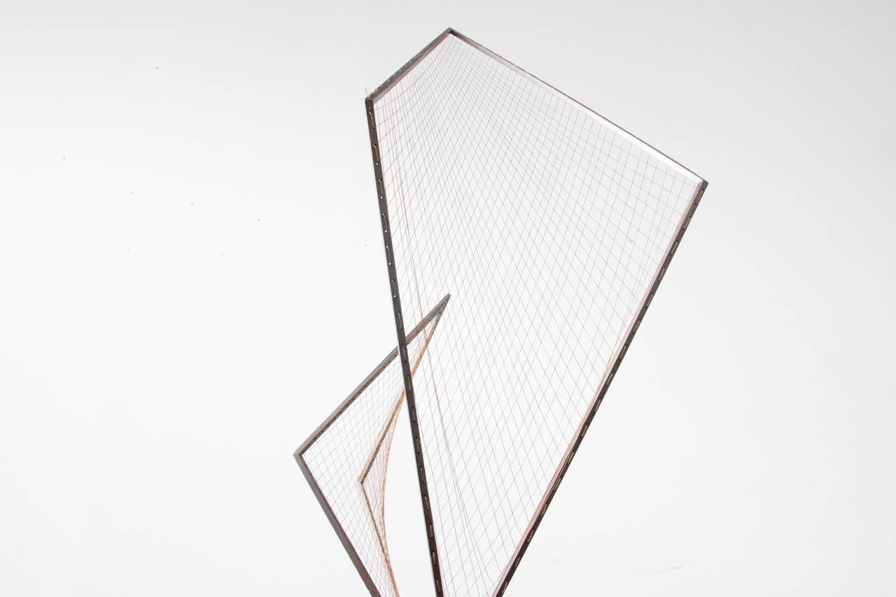 Ascending from a triangular prism base of iron, squared brass rods comprise the skeleton for a grid of strings. Fastened to perforations in the brass, each string and its bend contribute to the illusory quality of the curved grid.
