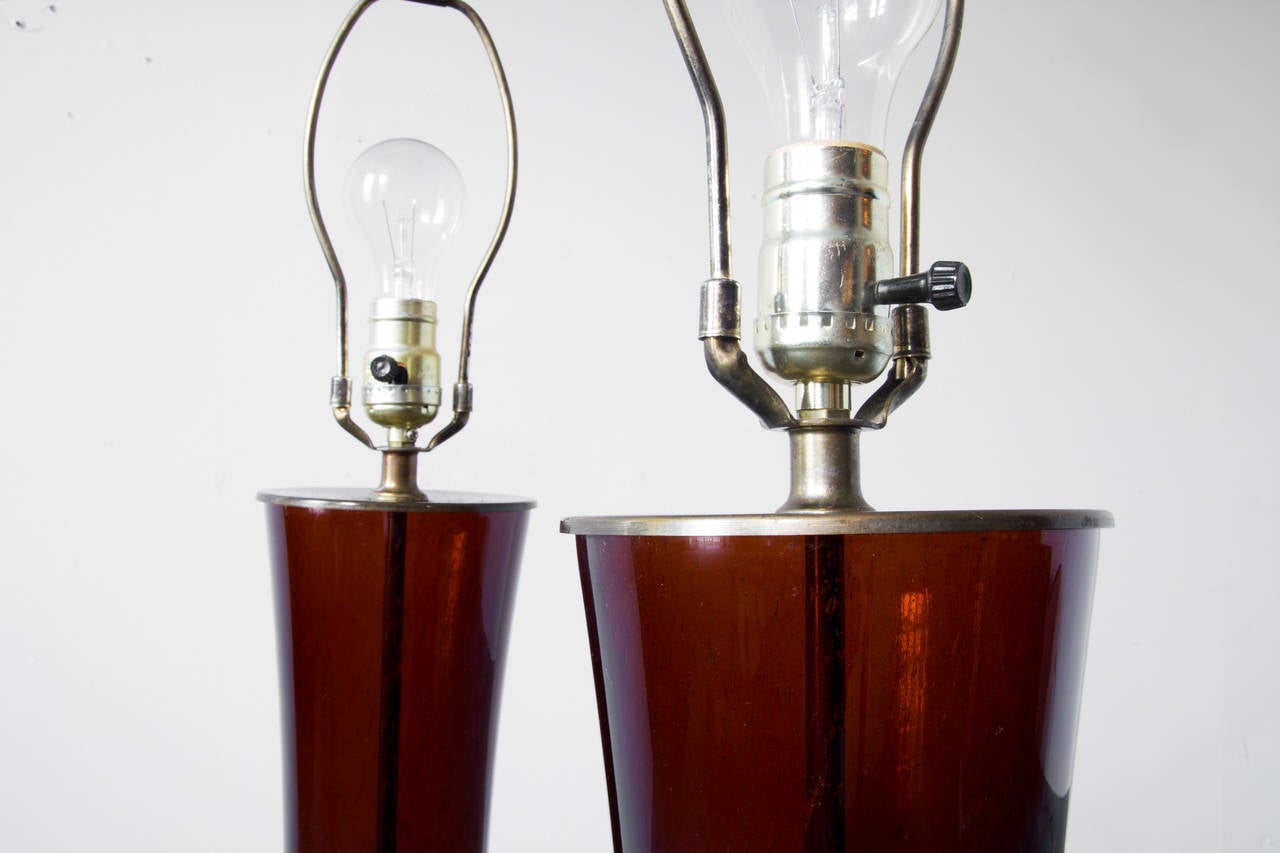 A pair of Italian lamps with bodies of dark amber glass capped by brass plates at top and bottom. Their hourglass shape contributes to their sleek and clean aesthetic. The turned brass finials are tall and ornamental.