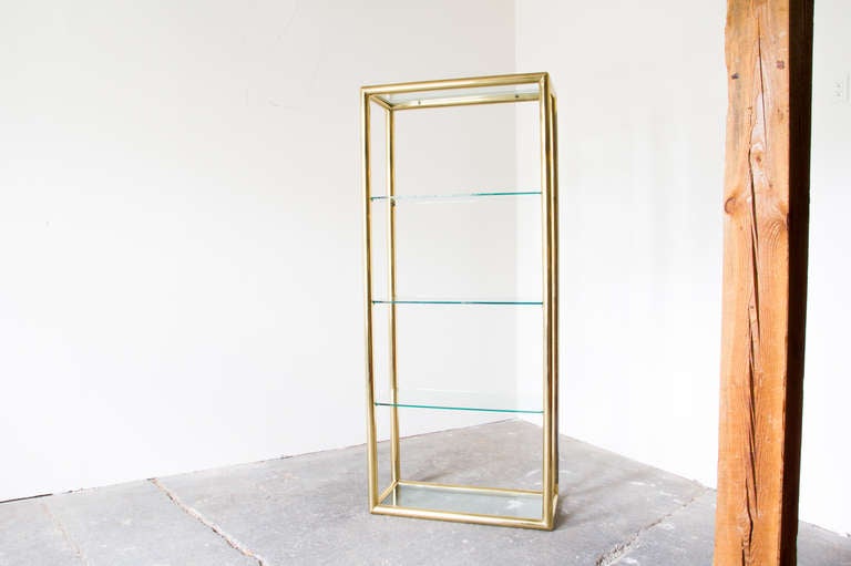 A shelving unit framed in brass with five shelves of thick, original glass. It is large, providing a good utility while retaining a delicate elegance. There is a shelf at ground level, as well as at the very top.