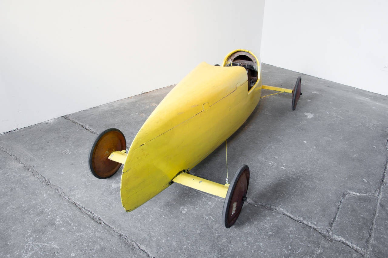 A sinuously carved and crafted, aerodynamic Soapbox Derby car in bright yellow. The steering still functions– controlled by a bent wire steering wheel in the cockpit. The frame is stacked solid bentwood, coming to a sharp point in the front. The