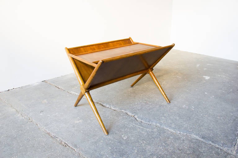 A functional magazine table in mahogany by T.H. Robsjohn-Gibbings for Widdicomb. The frame is comprised of tapered styles which cross-lap in the center forming an X on either side. There is a floating surface between two slanted surfaces for book