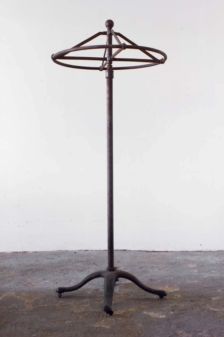 This iron garment rack rotates at the top, and rolls on four casters at the base. Though simple in form, it features a subtle, decorative relief on the support rails.