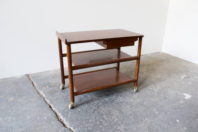 A rolling bar cart in walnut by William Pahlmann as part of his 1952 Hastings Square Collection, for Grand Rapids Bookcase & Chair Company. The drawer slides on a wooden track, and is accessible from either side. A lip on the end of the surface