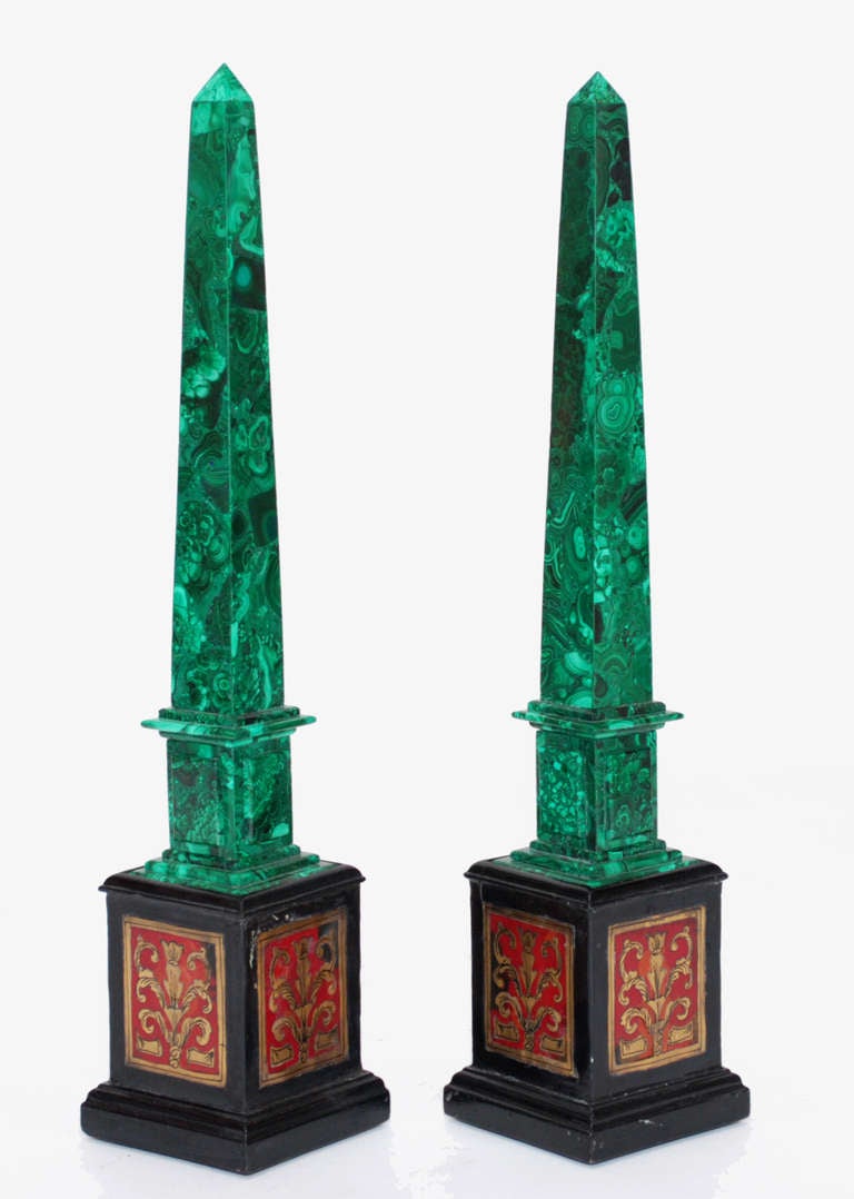Pair of gorgeous malachite obelisks on vintage 5.5” stands.  Each obelisk is raised on a square decorative wooden support with metal applique.  Above the malachite stepped platform base supports the square plinth with raised panels followed by the