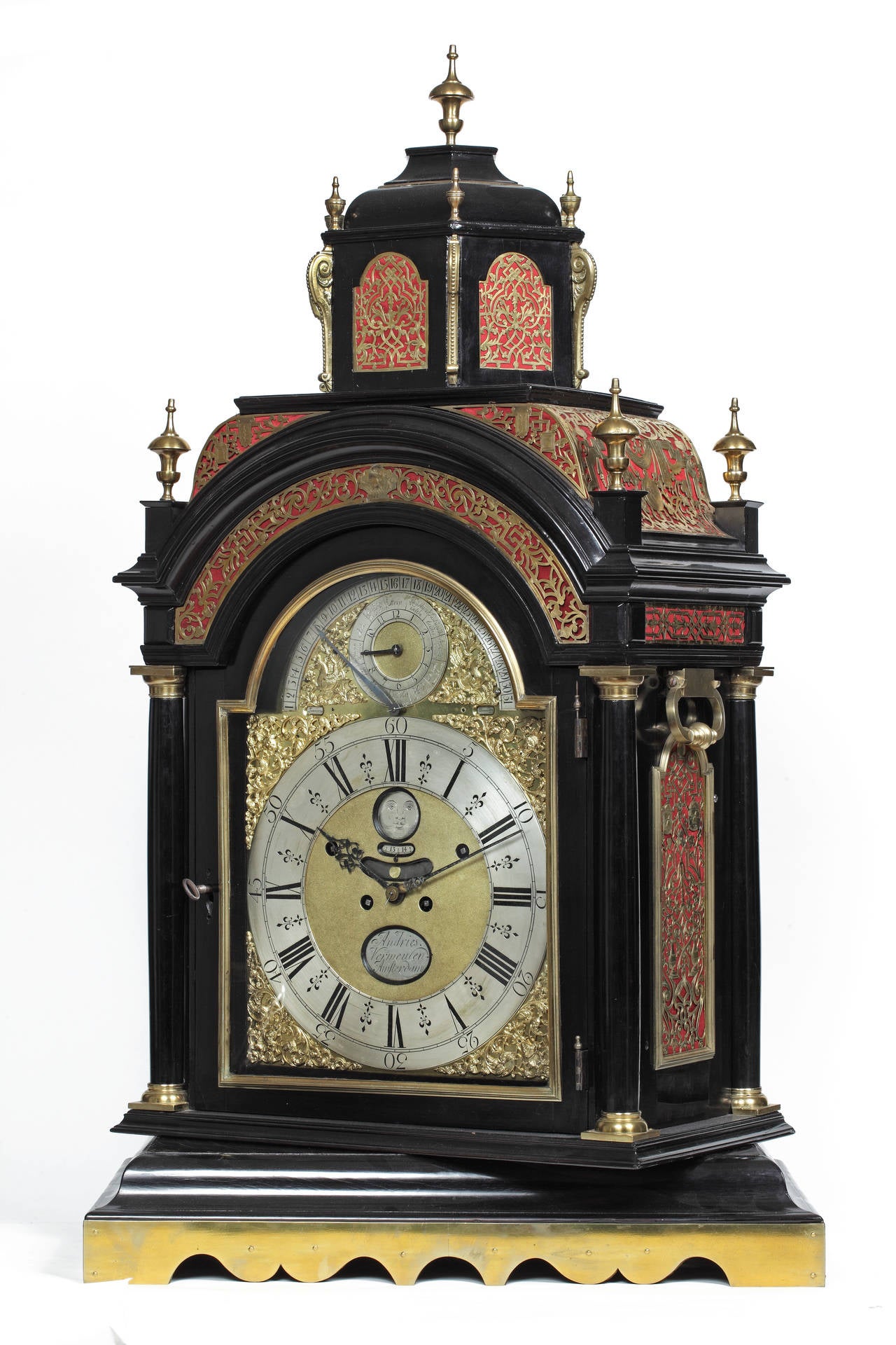 The ebony veneered case with cupola top and urn finial, with finely pierced and engraved brass sound frets against red ground to the top and sides, with finials and handles, on a waved turntable plinth. The dial with silvered Roman and Arabic