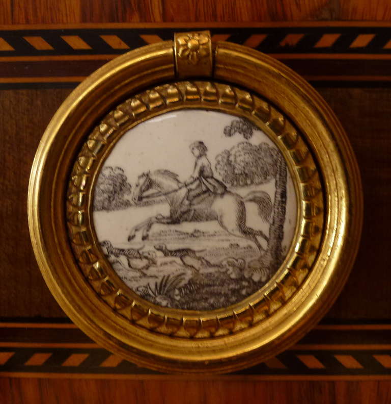 the top with a fan-shaped marquetterie in oval within a meander border, the drawer with ormolu ring-handles with white porcelaine plaquettes painted in black with horses and their riders, the sides also with te same plaquettes (one damaged), the