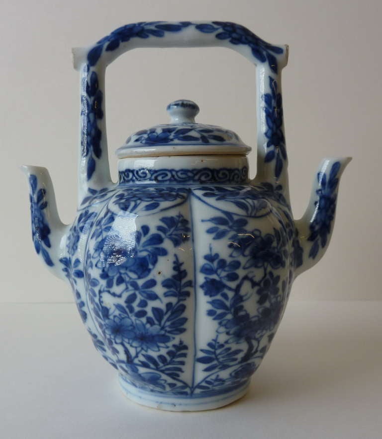 on footrim with two small curved spouts and stiff handle that consists of two vertical and 1 horizontal curved pieces; removable lid, inside the pot is divided in half by a straight wall; with blue floral decor in bedding on pleated wall and floral