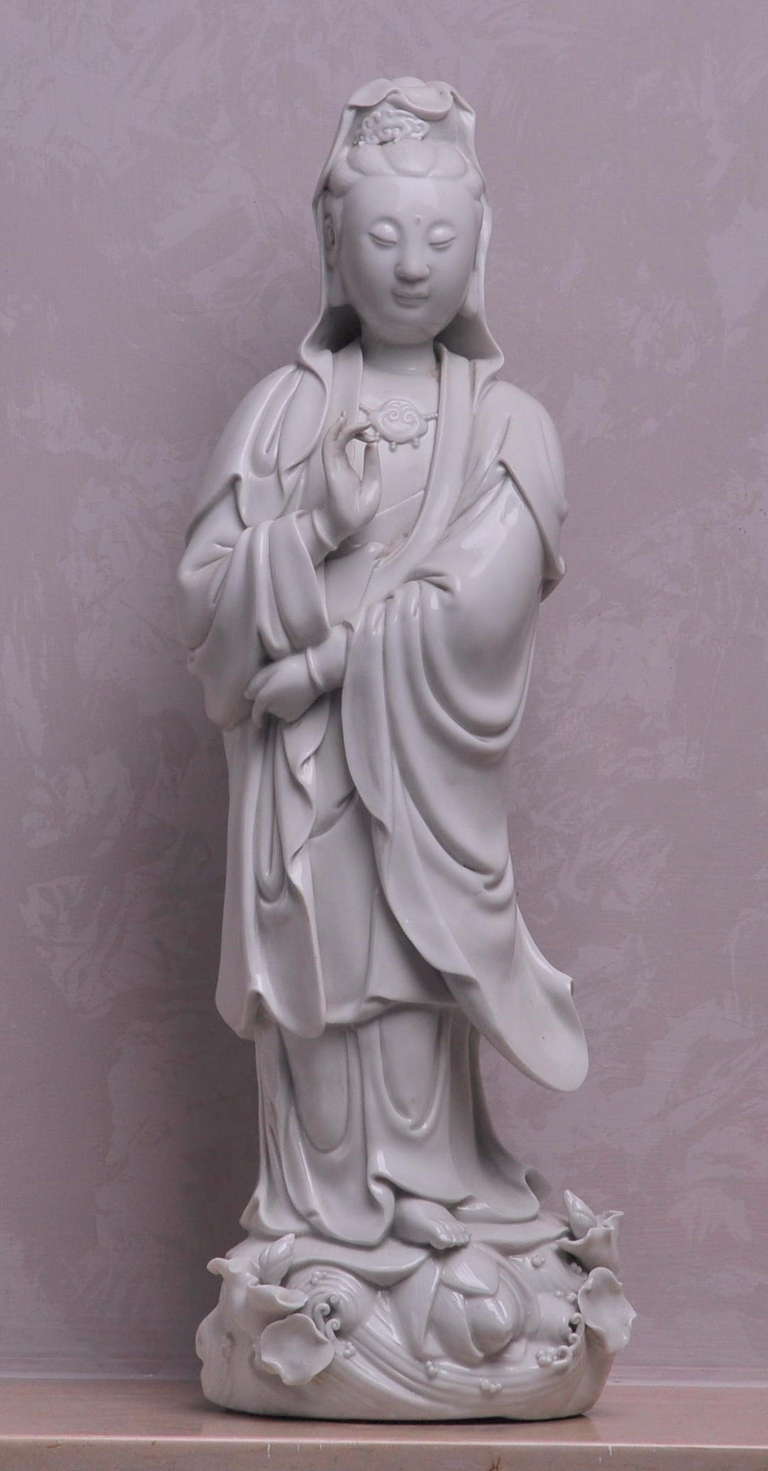 A BLANC-DE-CHINE FIGURE OF GUANYIN
QING DYNASTY,18TH CENTURY
standing on a the waves, the arms lifted to the waist, dressed in loose fitting robes opening at the chest to reveal a necklace and extending to a hood over the elaborate hair dress,