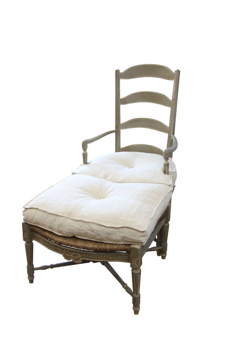 Provencal style armchair with its foot rest. The structure is made out of carved wood enhanced with a charming beige and grey patina. The rush seat dressed with thick natural-coloured cushions and the high ladder back add comfort to authenticity.