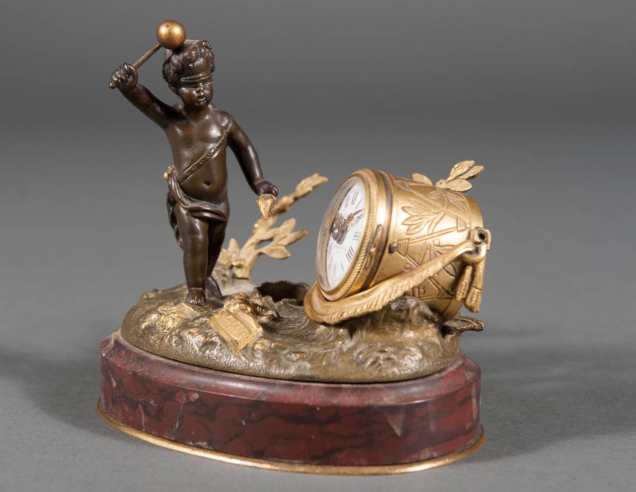 A charming miniature 19th century French gilt bronze patinated and Rouge marble clock cast with a blindfolded Cupid figure wielding a torch and drum mallet prepared to strike a drum-form clock, the circular white enamel dial with Roman hours and