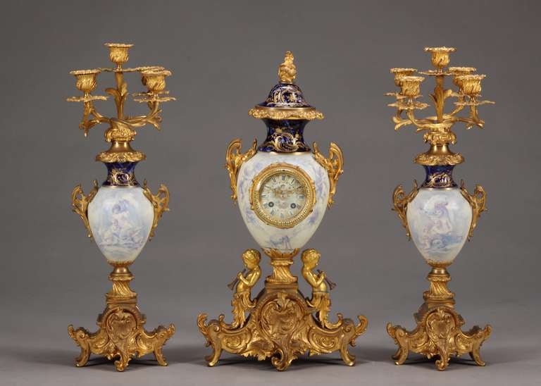 This excuisite 3-piece 19th century French ormolu mounted porcelain clock set comprising a central clock and a pair of flanking candelabra. The clock in urn shape form over a gilt bronze base surmounted with putti's playing a horn, and a painted