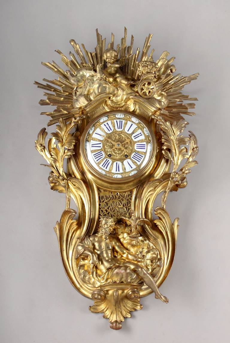 A large French Louis XV style gilt bronze cartel clock.
After the model by Jacques Caffièri.
The cartouche-shaped case representing sunrise and sunset, surmounted by a putto and doves in the Venus’ chariot above clouds in front of a large sunburst.