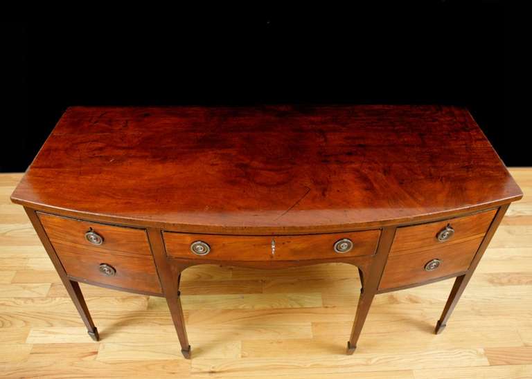 A handsome George III sideboard in mahogany with bowed front and square tapered legs ending in spade feet, with central frieze drawer flanked by two cellarette drawers. Top is composed of a single solid plank of mahogany, England, circa
