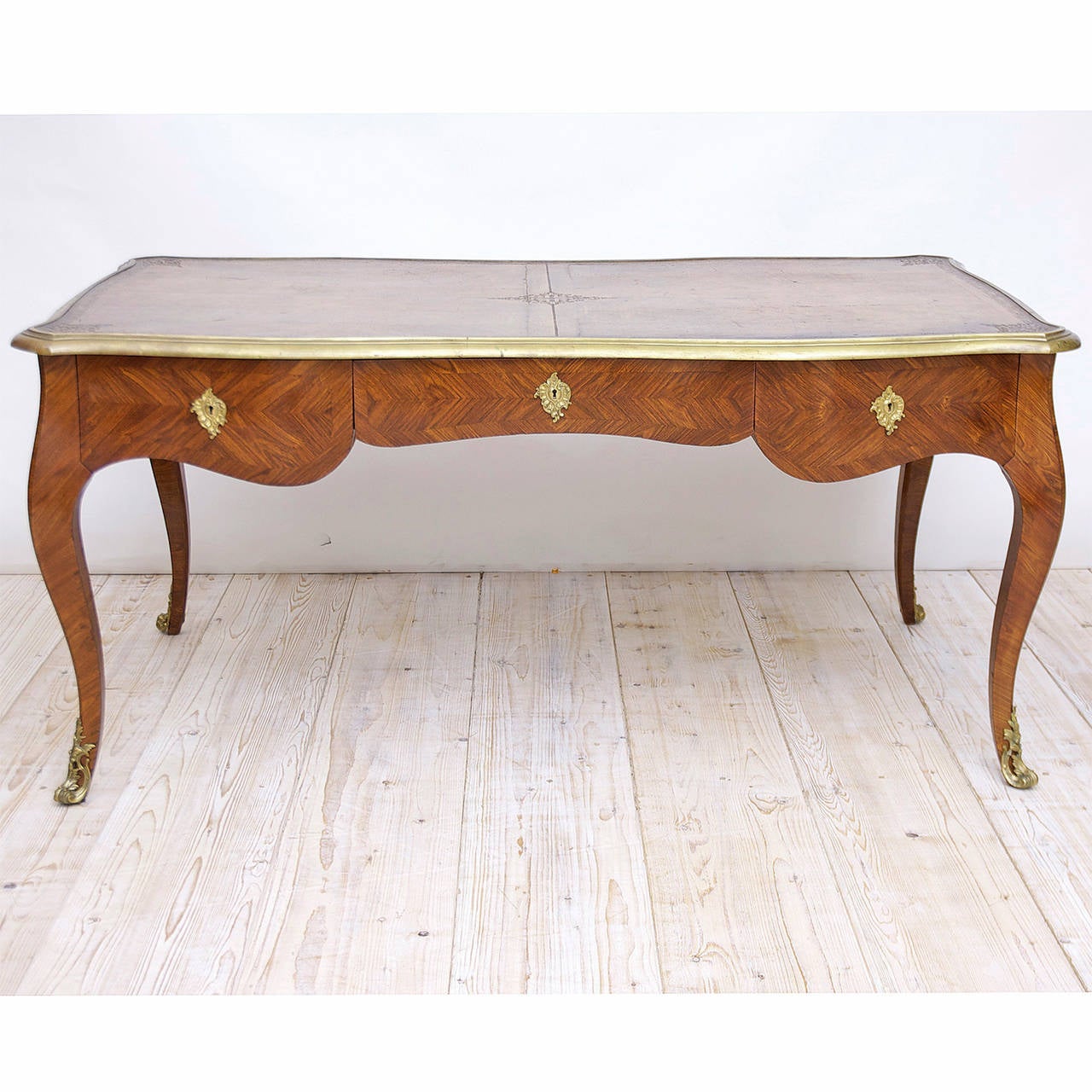 An exceptionally fine bureau plat or desk in the Louis XV style in parquetry kingwood with serpentine form on all four sides, cabriole legs terminating with bronze doré sabots, stepped molding in brass around perimeter of inset leather top. Offers