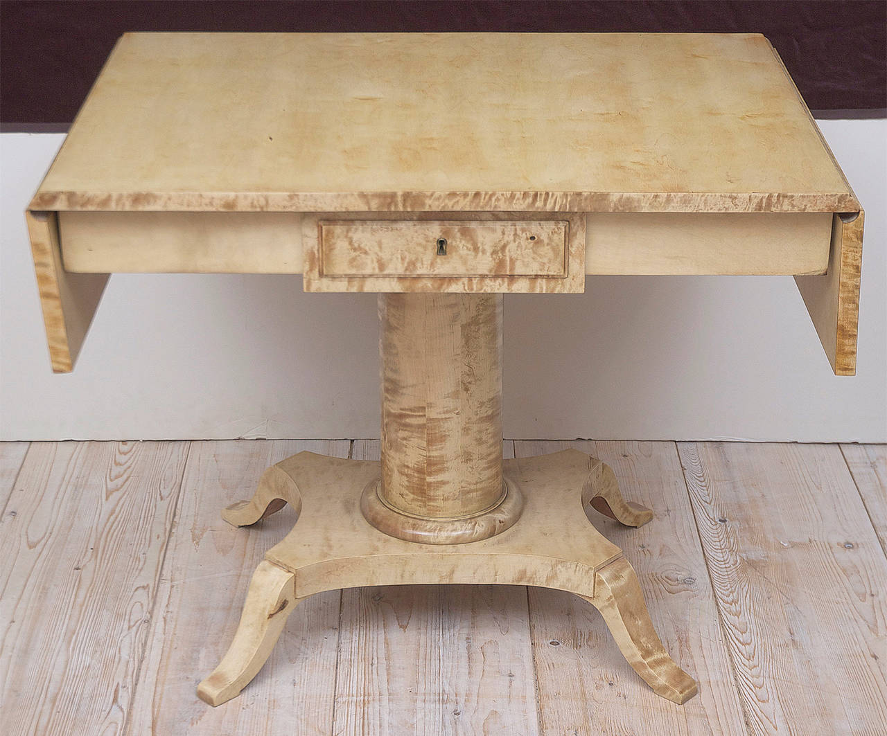 A very lovely table in ivory-toned birchwood with drop-leaves on cylindrical column resting on quarter-form base with saber feet. Offers one-drawer. Traditionally used in front of a sofa and offering a multipurpose table for tea, games or writing,