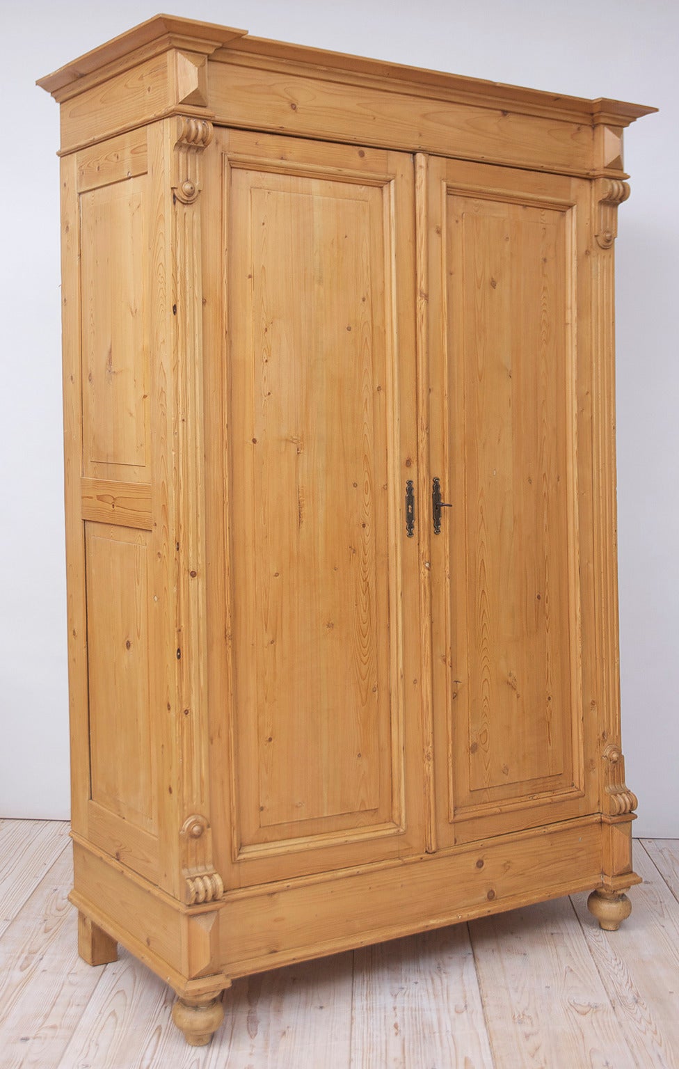 A pine armoire with two raised-paneled doors, carved escutcheons defining the corners, side panels and resting on turned bun feet. Offers interior shelving. Dating from the rise of the German Republic (Gründerzeit period). Germany, circa