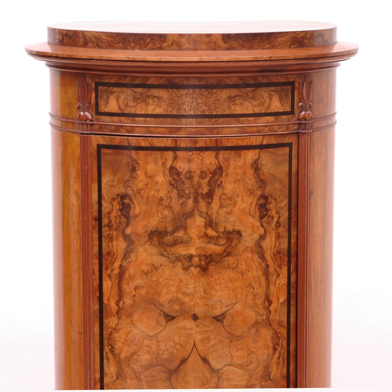 A fine pedestal cabinet whose cylindrical form is enhanced by ebonized details and by the play of highly figured bookmatched walnut veneers. This Empire or Biedermeier cabinet is an object of singular beauty that offers a place to exhibit a marble